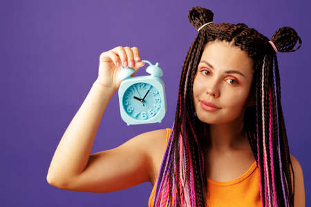 Young stylish woman with braids holding blue alarm clock, time concept