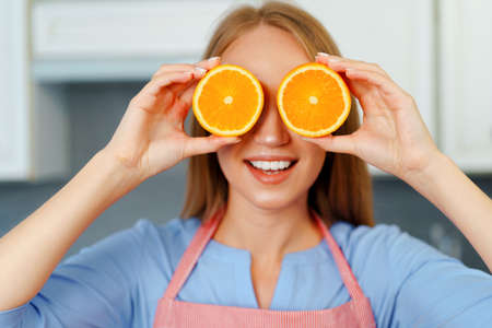 Beautiful caucasian woman in red apron holding ripe oranges while standing in kitchenの写真素材