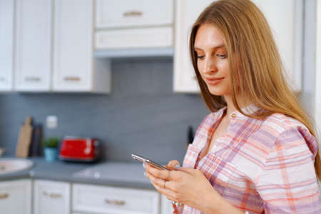 Pretty young woman in casual clothes using her smartphone in kitchen