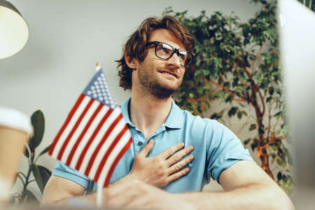 Young bearded businessman sitting at table with laptop and american flagの写真素材