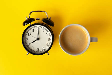 Alarm clock and coffee cup on yellow background, top view