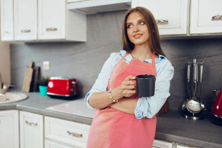 Pretty young woman in red apron enjoying a cup of coffee in her kitchenの写真素材