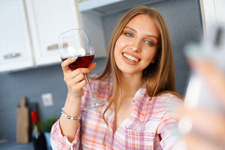 Happy relaxed young woman standing in kitchen with glass of red wine and using her smartphoneの写真素材