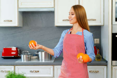 Beautiful caucasian woman in red apron holding ripe oranges while standing in kitchenの写真素材