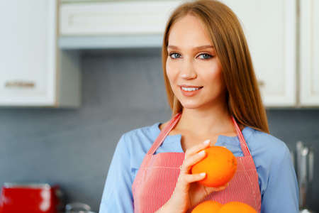 Beautiful caucasian woman in red apron holding ripe oranges while standing in kitchenの写真素材