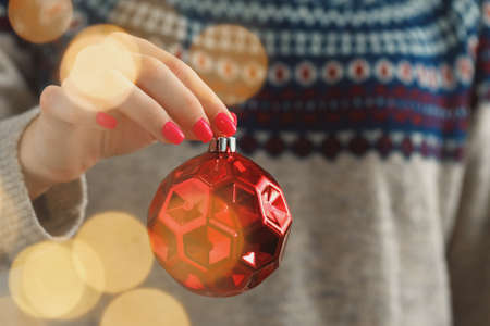 Hand of a woman in festive sweater holding Christmas bauble with blurred lightsの写真素材