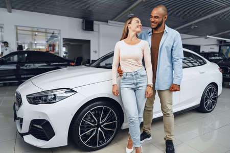 Happy couple of caucasian woman and african american man standing near their new car inside car salonの写真素材