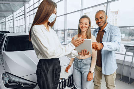 Car saleswoman wearing medical mask shows buyers couple something on digital tabletの写真素材
