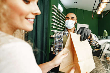 Male coffee shop worker giving ready order to the client wearing face mask
