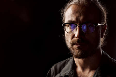 Dark studio portrait of a middle-aged man with beard in glasses