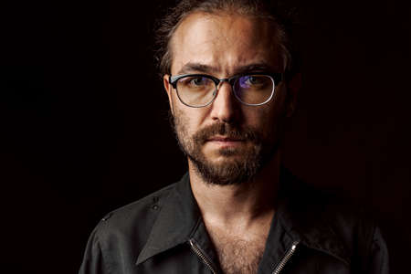 Dark studio portrait of a middle-aged man with beard in glasses