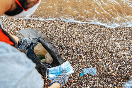 Man volunteer collecting used medical masks on the beach near the ocean