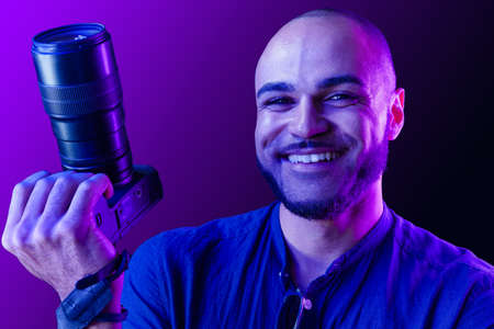 Black man with camera standing against purple background in neon light