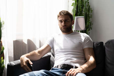 Sad thoughtful bearded man sitting on a sofa near window
