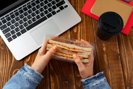 Hands of a woman holding sandwich above working table with laptopの写真素材