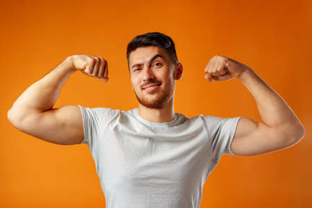 Positive smiling young man showing his strong arms against yellow background