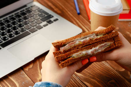 Hands of a woman holding sandwich above working table with laptopの写真素材