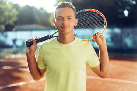 Young man teenager with tennis racket standing near net on clay courtの写真素材