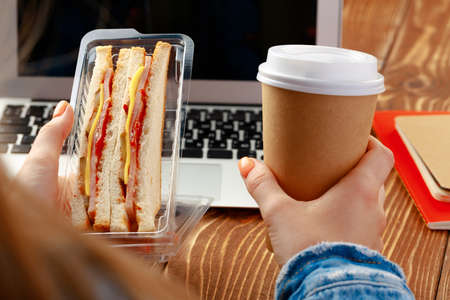 Hands of a woman holding sandwich above working table with laptopの写真素材