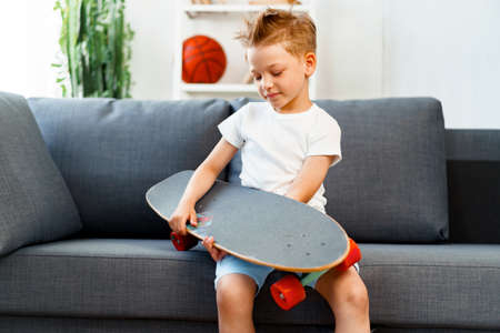 Little boy sitting on sofa at home and holding skateboard
