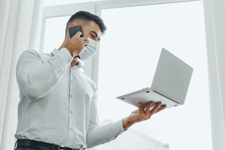 Handsome man businessman in medical mask using his laptop while talking on his cellphoneの写真素材