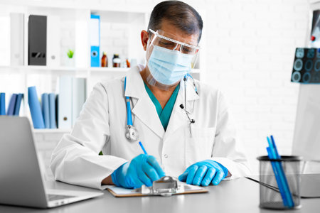 Middle-aged male doctor in face shield and mask sitting at the table at his office