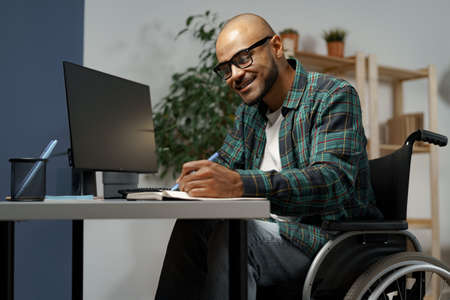 Disabled young african american man in wheelchair using computer while sitting at his working tableの写真素材