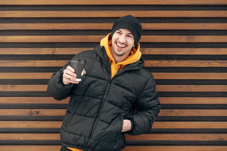Young man in winter clothes standing with coffee to go in a street against wooden wallの写真素材