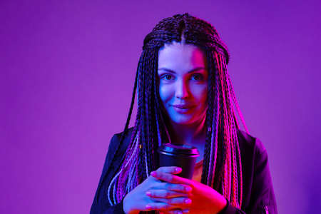 Portrait of a woman with long afro braids holding a cup of coffee against purple neon background
