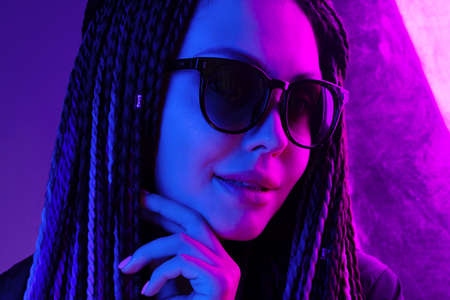 Stylish young woman with braids wearing sunglasses against purple background