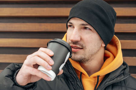 Young man in winter clothes standing with coffee to go in a street against wooden wall