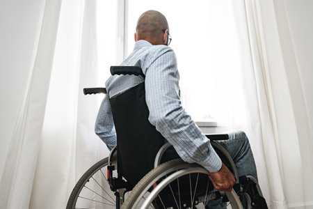 African american man in wheelchair sitting near the window alone at homeの写真素材