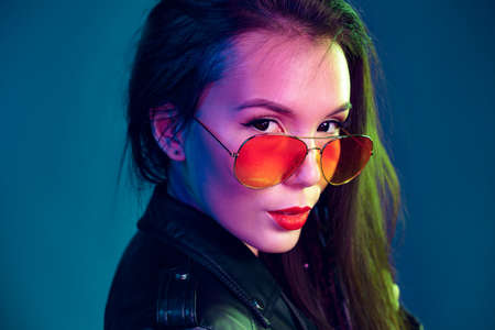 Asian girl in sunglasses and black leather jacket posing against dark blue background