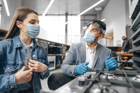 Male salesman in hypermarket describes features of gas stove to female client, both wearing face masksの写真素材