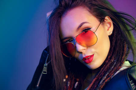 Asian girl in sunglasses and black leather jacket posing against dark blue background