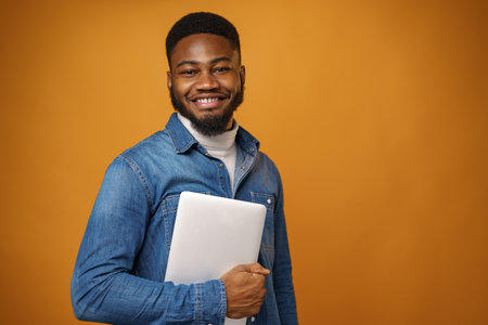 Young african american man standing with laptop against yellow backgroundの写真素材