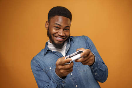Young african american man holding joystick playing video games, yellow background