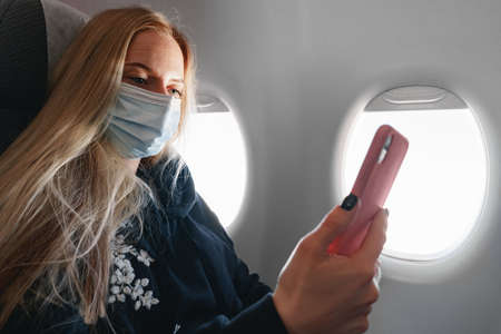 Young woman with long hair sitting in plane by the window and using smartphone
