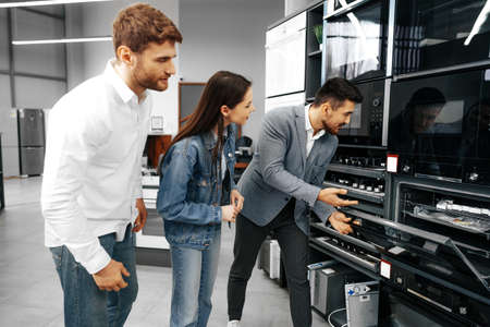 Male shop assistant helps young couple to choose new home appliance