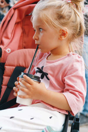Blonde toddler girl sitting in the stroller and drinking from disposable cupの写真素材