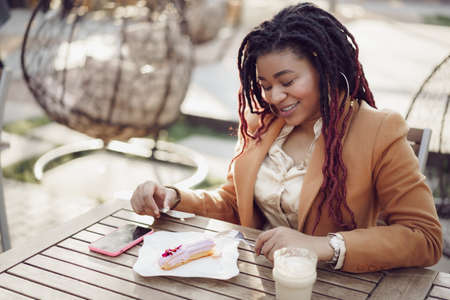 Smiling african american woman drinking coffee and eating dessert in outdoor cafeの写真素材