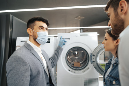 Salesman in hypermarket wearing medical mask demonstrates his clients a new washing machine