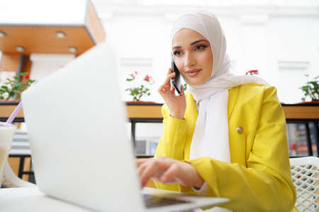 Young muslim businesswoman in headscarf sitting in cafe and talking on the phoneの写真素材