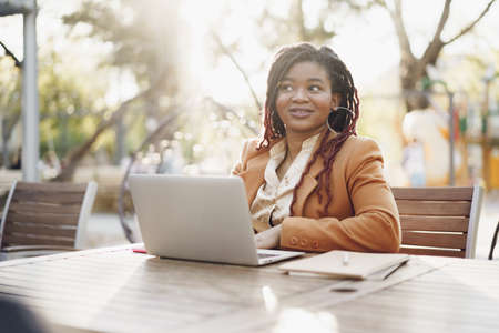 Young smiling african american woman sitting at the table in street cafe and using laptopの写真素材