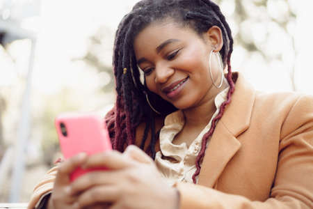 Young african american woman sitting in street cafe and holding smartphone in her handsの写真素材