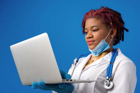 Young african american woman doctor using laptop against blue backgroundの写真素材