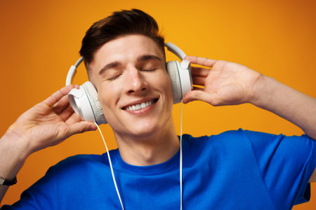Young man in blue t-shirt listening to music with headphones against yellow backgroundの写真素材
