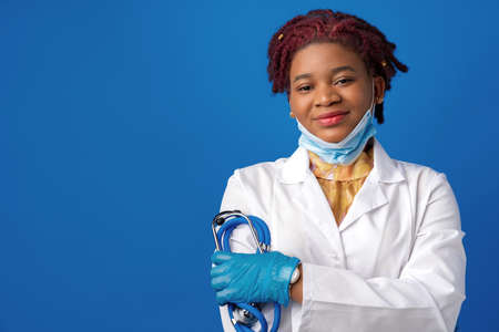 Portrait of african female doctor in lab coat with face mask and stethoscope against blue backgroundの写真素材