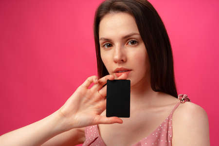 Attractive smiling young woman holding black credit card against pink studio background