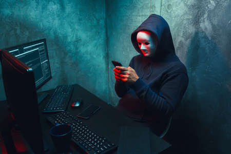 Anonymous hacker wearing face mask working on computer in dark roomの写真素材